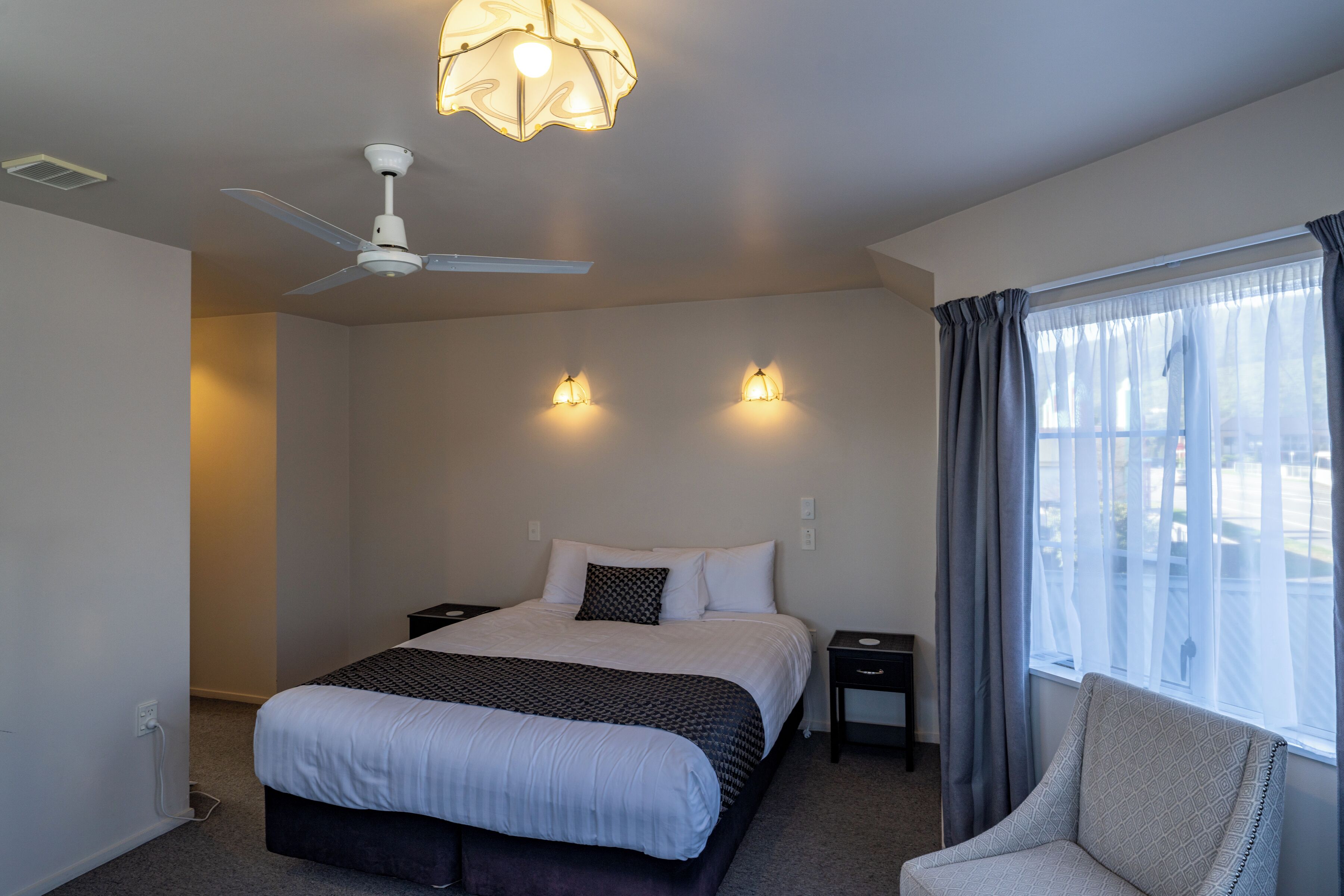 A hotel room with a bed, a ceiling fan, two bedside lamps, a window with curtains, and a chair.