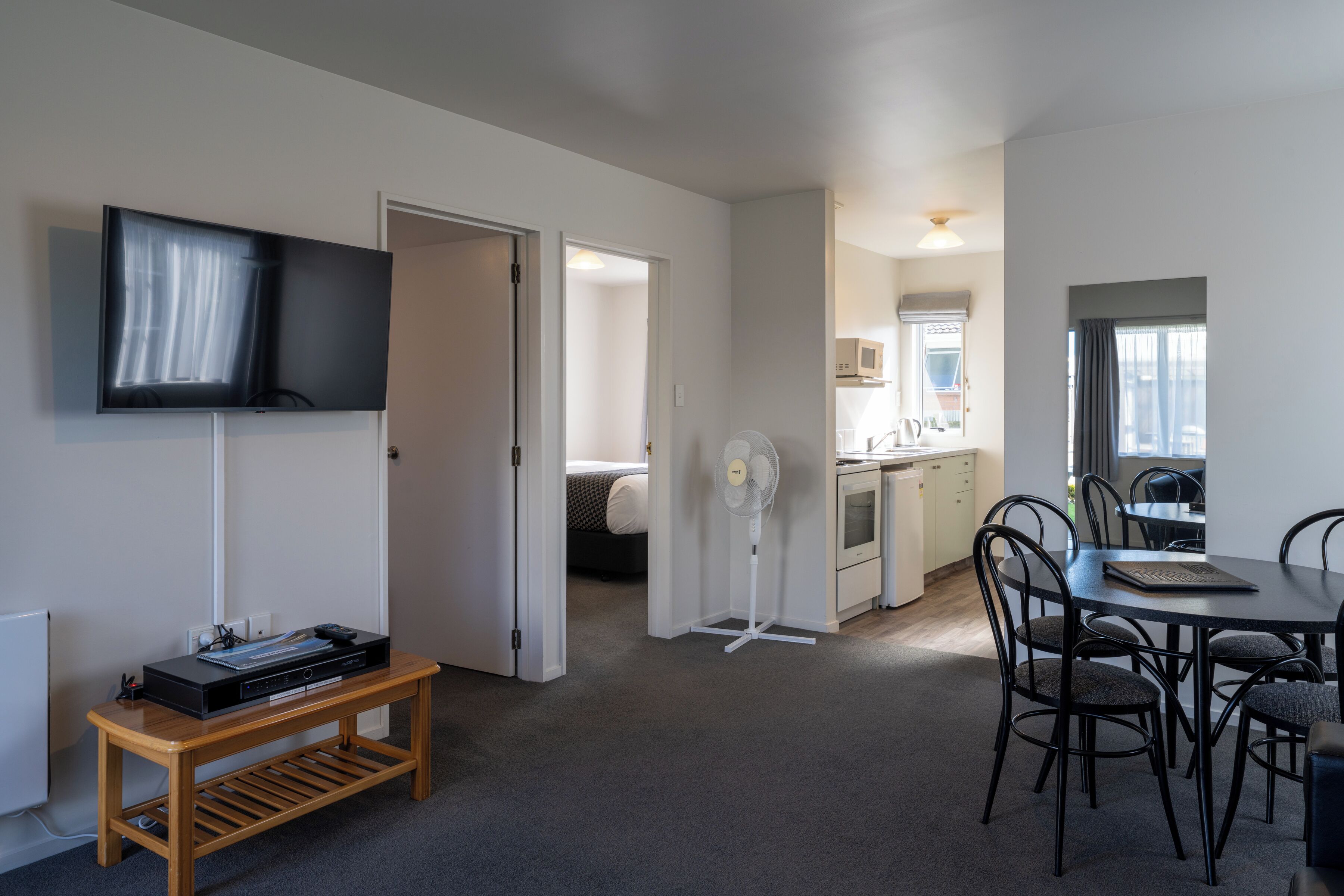 Una habitación moderna y luminosa con una televisión de pantalla plana, un banco de madera, una mesa de comedor con sillas, una kitchenette y una cama al fondo.