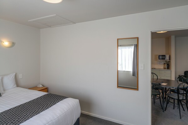 A hotel room with a bed, bedside table, and a mirror. There is a kitchenette with a microwave and a dining area with chairs.