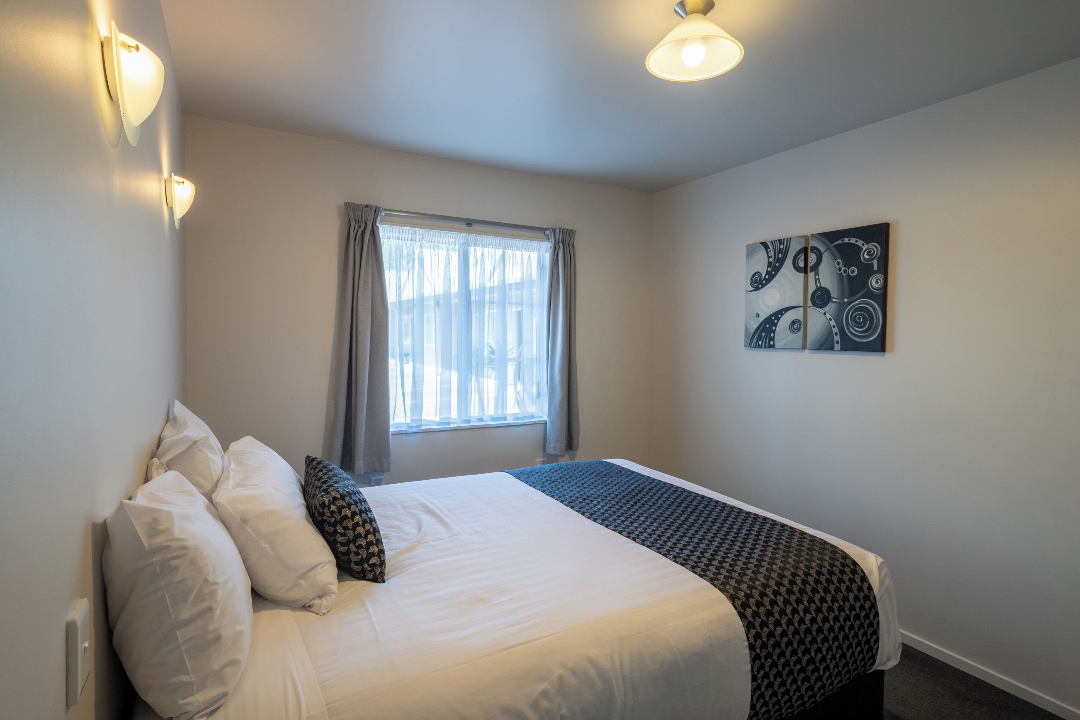 A neatly made bed with white and patterned bedding, a window with curtains, and a wall-mounted abstract art piece.