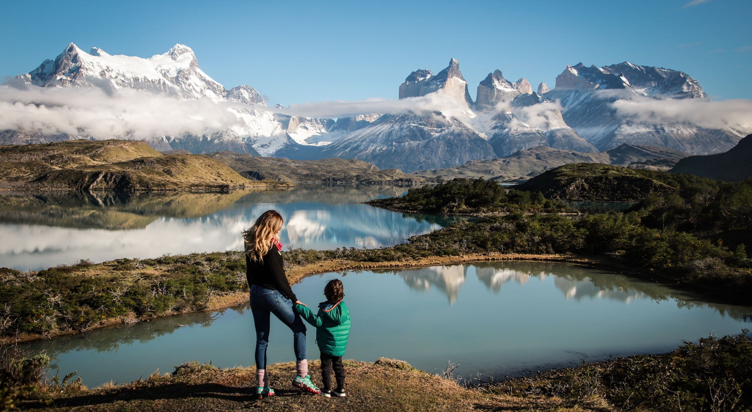 Un lac avec des montagnes en arrière-plan et une personne debout au bord de l’eau.