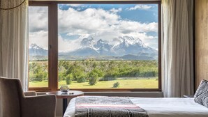 A hotel room with a large window offering a mountain view, a bed, a chair, and a small table with a fishbowl. - Rio Serrano Hotel + Spa (Torres del Paine)