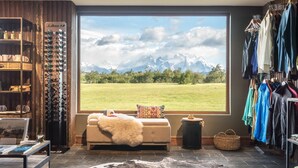 A room with a large window overlooking a mountain landscape, a fur throw on a bench, a clothing rack, and shelves with various items. - Rio Serrano Hotel + Spa (Torres del Paine)