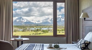 A hotel room with a large window offering a mountain view, a bed with a patterned bedspread, a bedside table with a book and a cup, and a lamp on the wall.