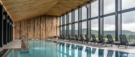 An indoor swimming pool with wooden walls and large windows offering a view of the hills.