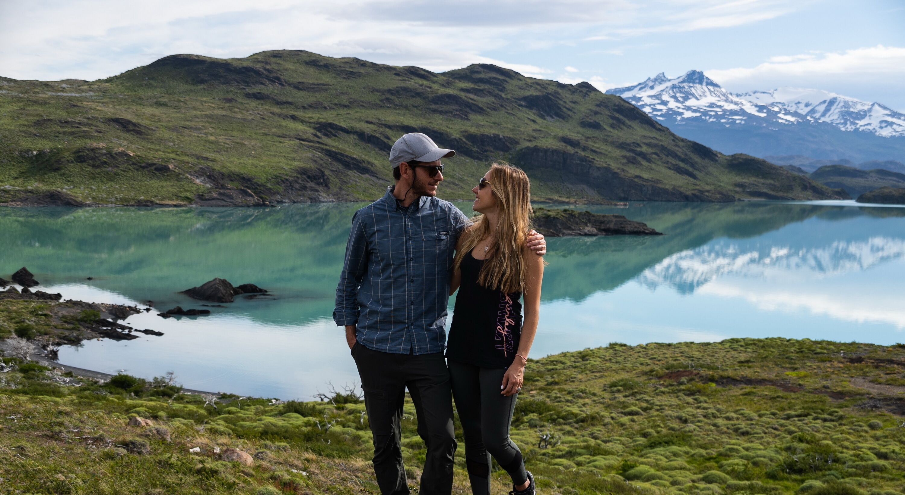 Una pareja de pie en una colina cubierta de hierba, con vistas a un lago y montañas al fondo.