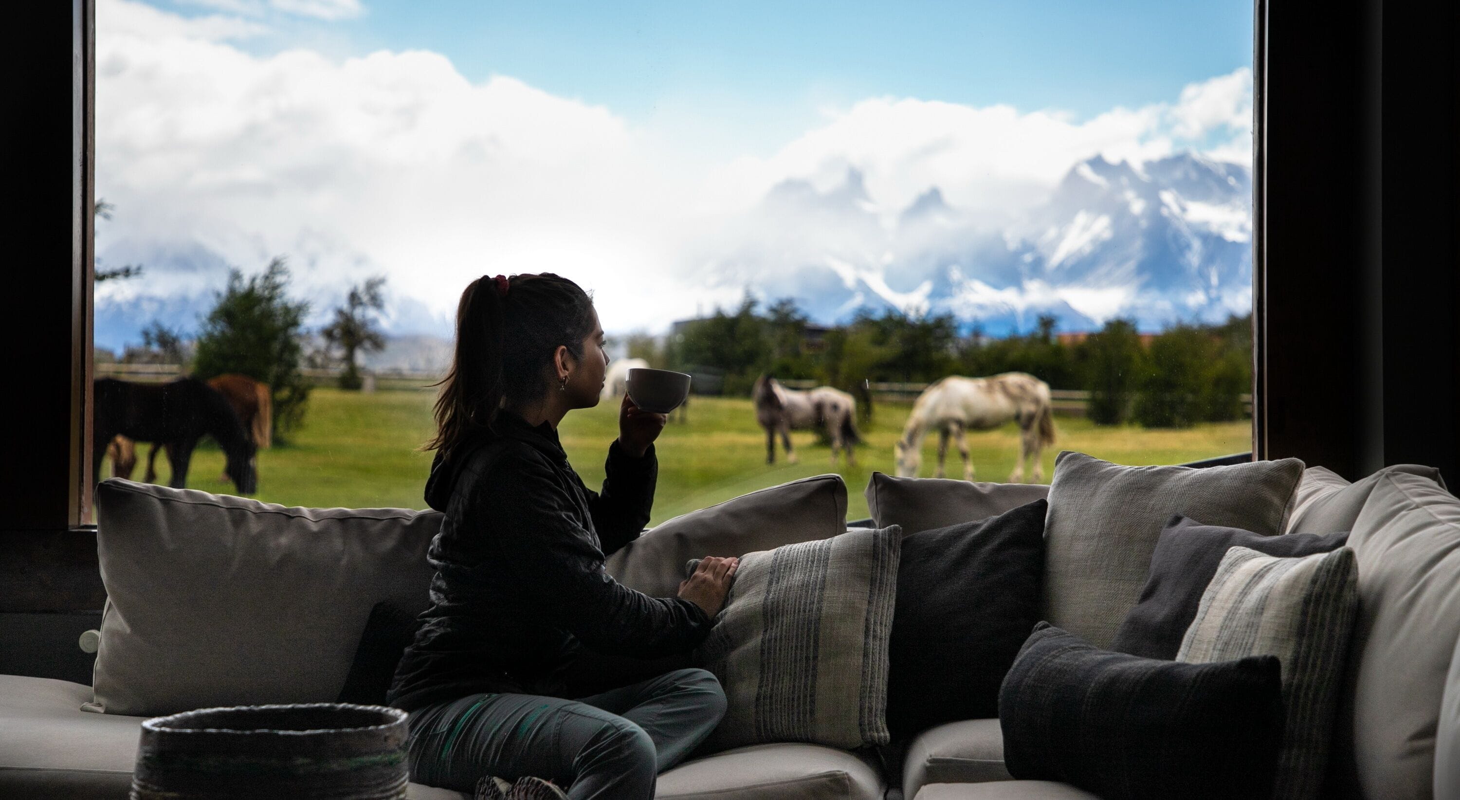 A person sitting on a couch with a view of horses grazing in a field and mountains in the background.