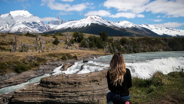 Eine Person sitzt auf einem Felsen mit Blick auf einen Fluss, einen Wasserfall und verschneite Berge im Hintergrund.