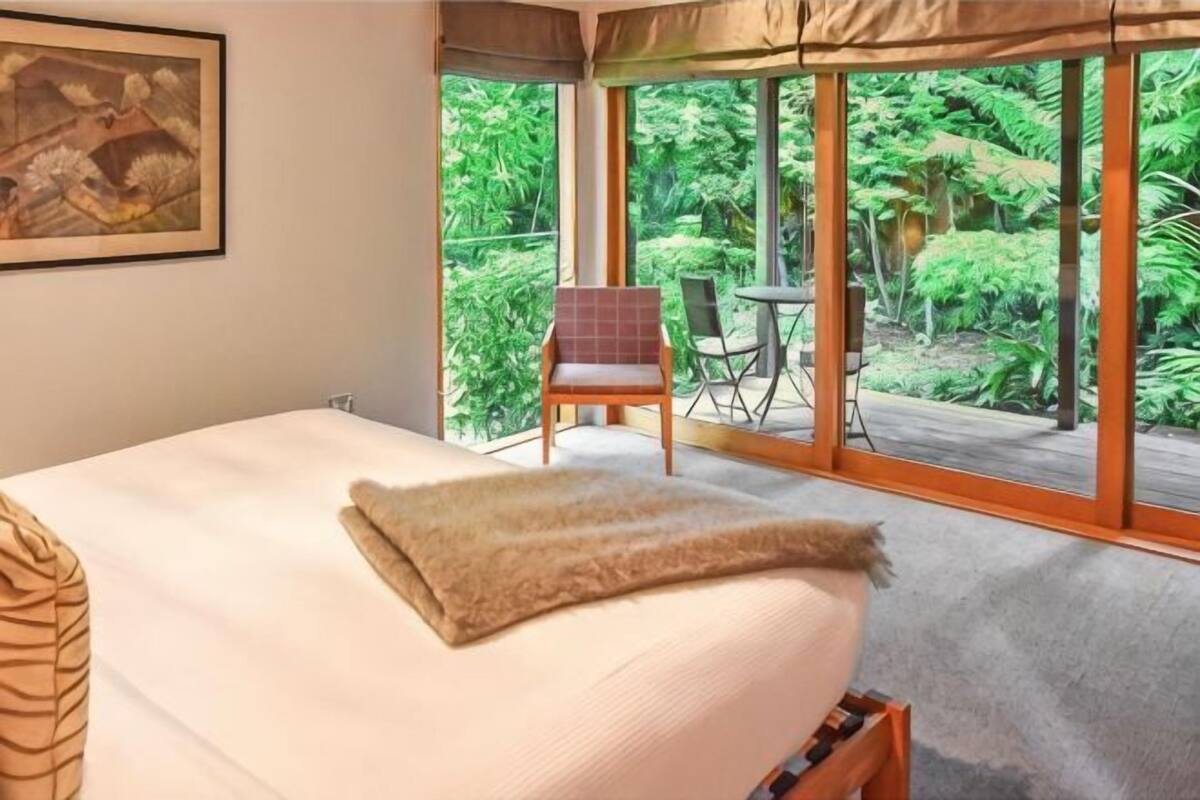 Un dormitorio con una cama grande, una silla, una mesita y vistas a un verdor exuberante a través de amplias ventanas.
