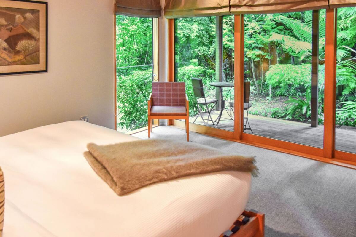 Un dormitorio con una cama grande, una silla y vistas a un jardín exuberante a través de amplios ventanales.