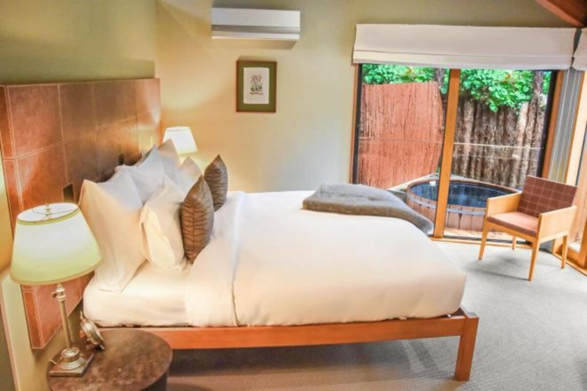 A bedroom with a bed, bedside lamps, a chair, and a view of a hot tub.