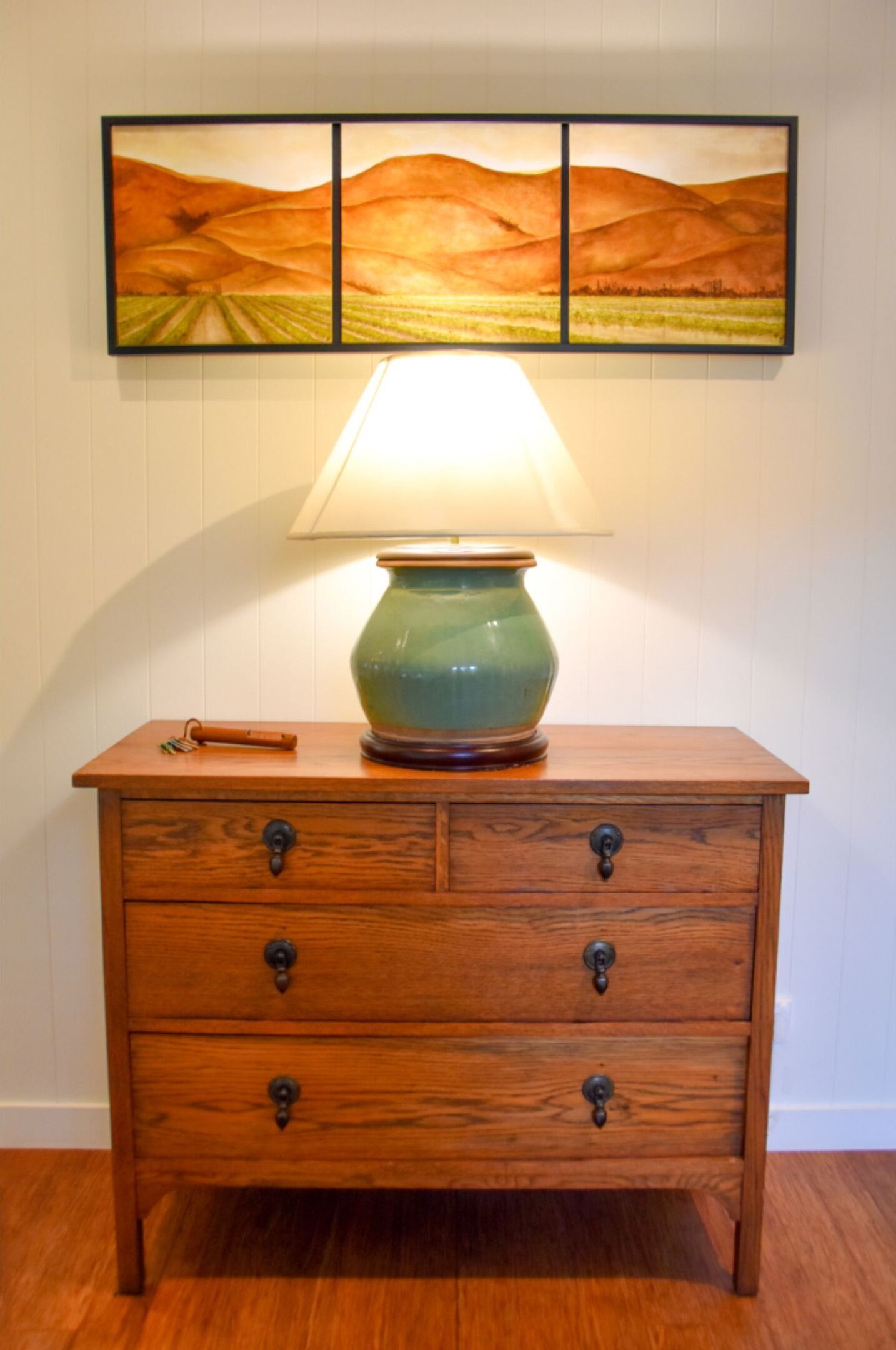 Una mesita de noche de madera con una lámpara verde, un cajón y una imagen encima.