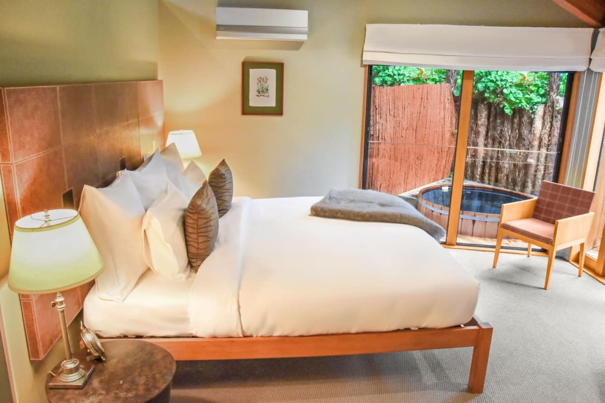 Un dormitorio con una cama grande, una mesita de noche con lámpara, una silla y vistas a una valla de madera y vegetación al exterior.