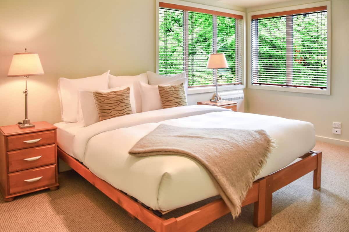 Una cama bien hecha con sábanas blancas y una manta color beige, flanqueada por dos lámparas de noche, en una habitación con ventanas amplias y una mesita de noche de madera.