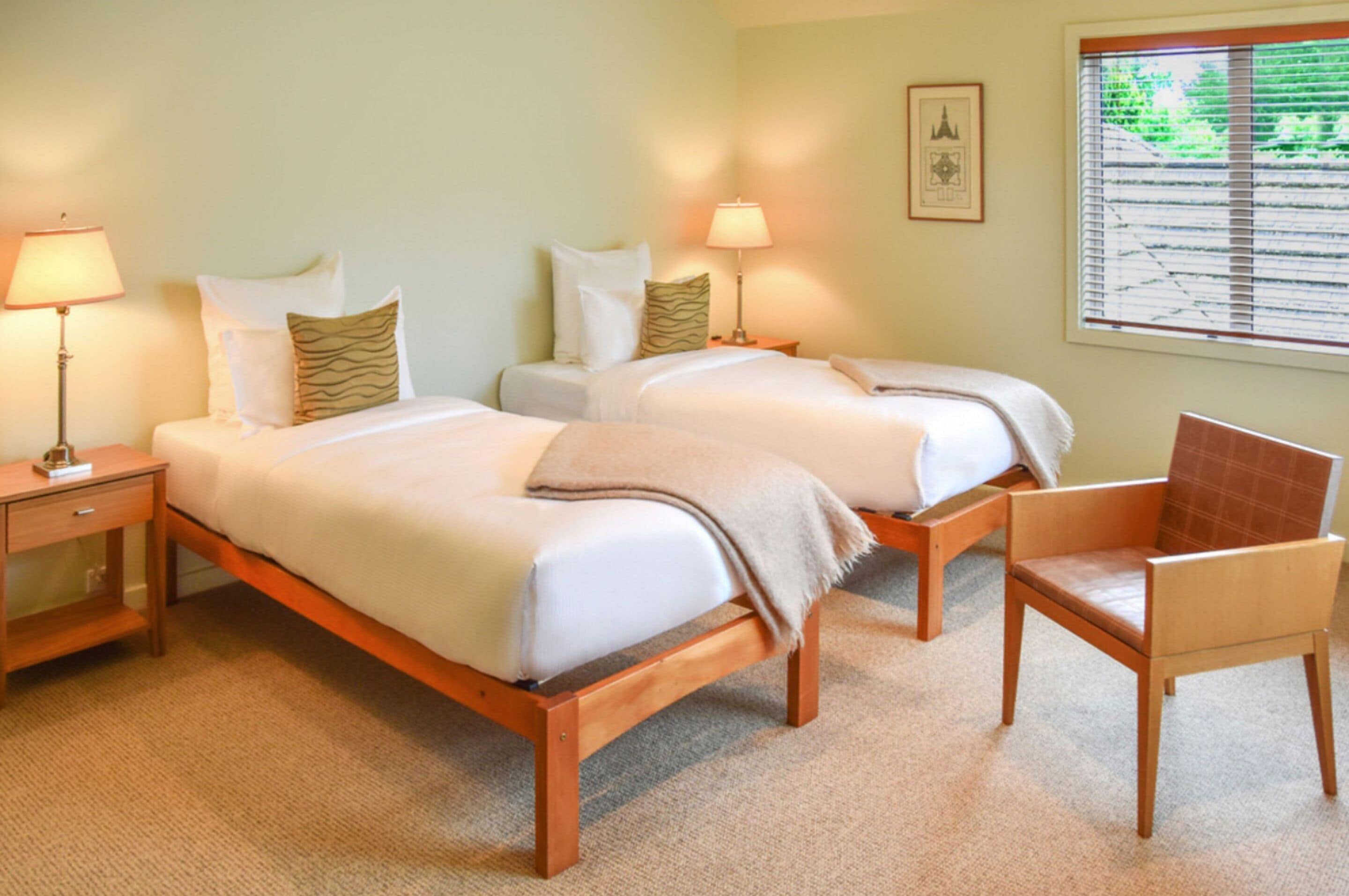 Une chambre d’hôtel avec deux lits, une chaise, une table de chevet et une fenêtre avec des stores.