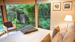 A bedroom with a large bed, a chair, and a view of a lush forest through large windows. - River Birches (Turangi)