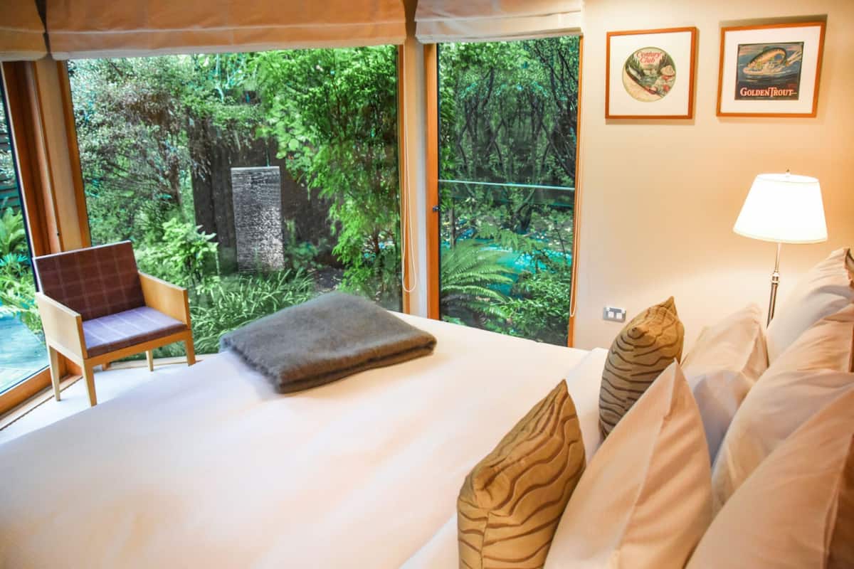 Un dormitorio con una cama grande, una silla y vistas a un bosque frondoso a través de amplias ventanas.
