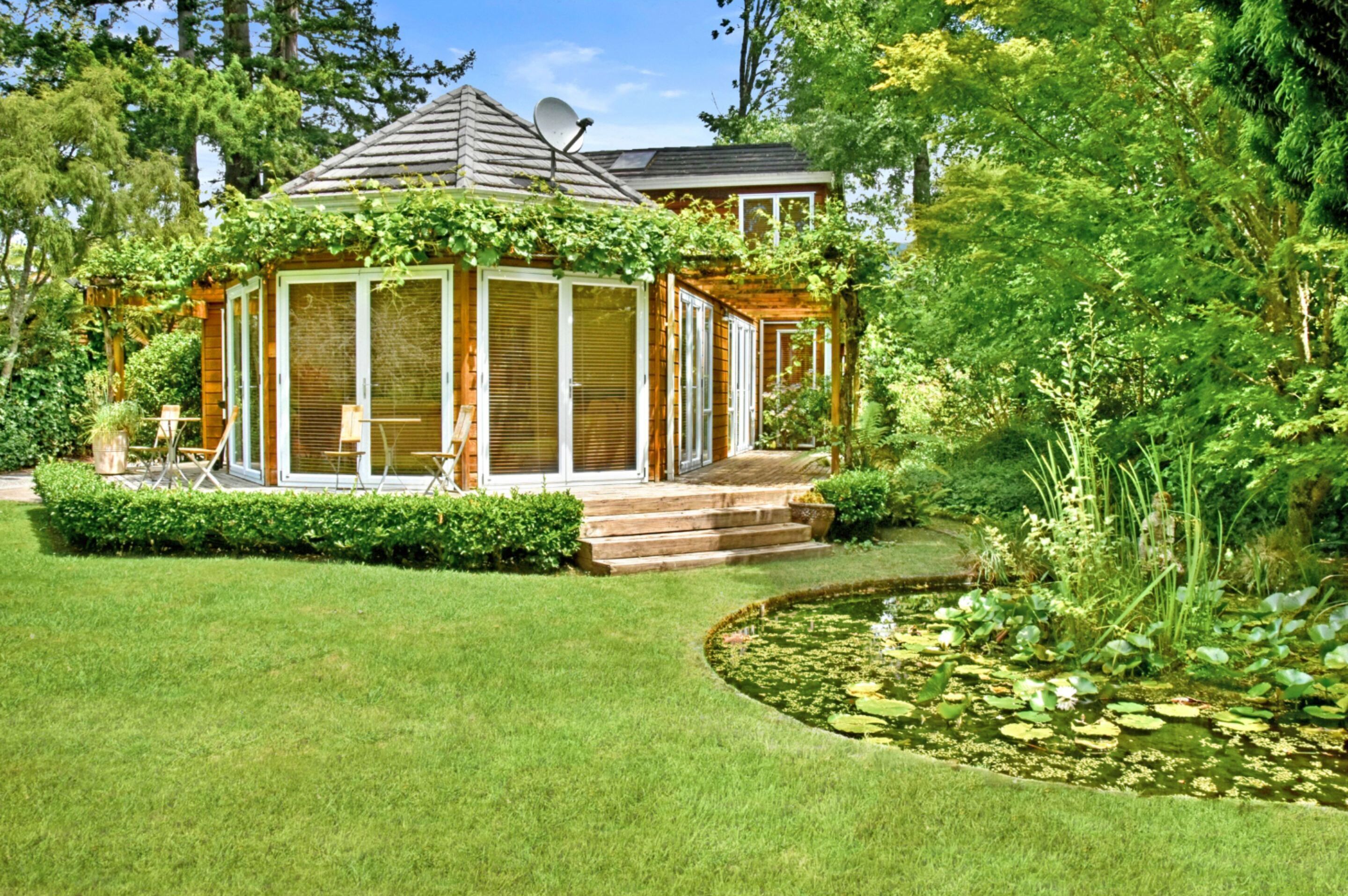 A wooden gazebo with a glass enclosure, surrounded by a well-manicured lawn and a pond with lily pads.