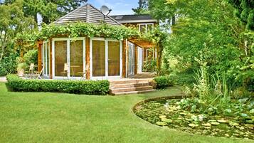 A wooden gazebo with a glass enclosure, surrounded by a well-manicured lawn and a pond with lily pads.