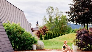 Une piscine avec une personne assise au bord, entourée de verdure et d’un bâtiment doté d’un toit en pente.