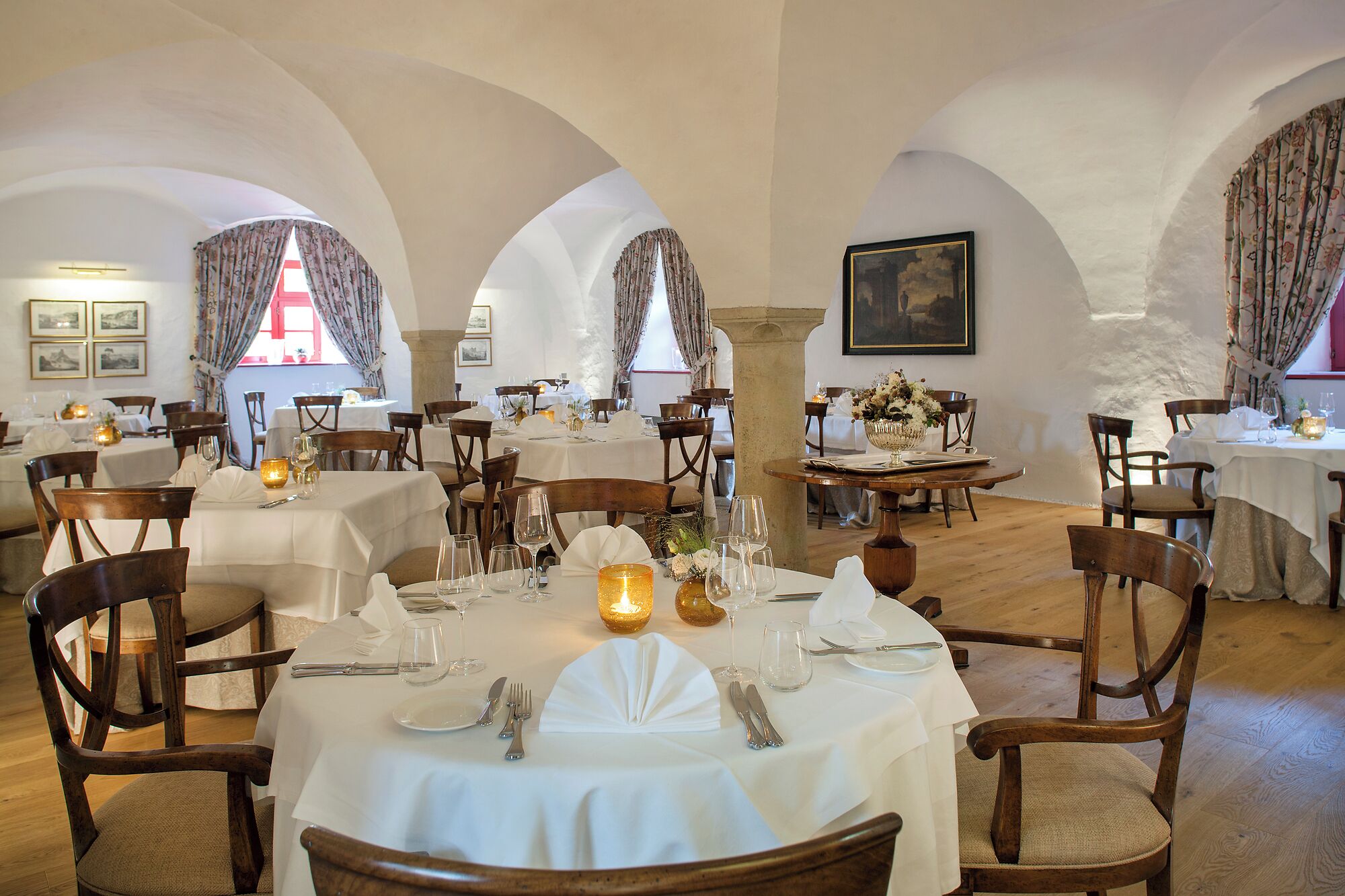 Un restaurant doté de nombreuses tables dressées pour le service, avec des nappes blanches, des chaises en bois et des centres de table illuminés par des bougies.