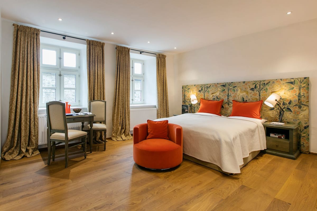 A bedroom with a bed, two chairs, a small orange ottoman, a nightstand with a lamp, and a window with curtains.