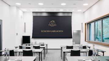 Una sala de conferencias con una pantalla grande que muestra 'TAURORA SCHLOSS GABELHOFEN'. Las mesas están dispuestas con sillas y botellas de agua.