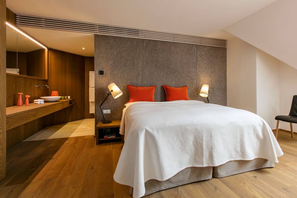 A modern bedroom with a large bed, two bedside lamps, a wooden nightstand, a mirror, and a small seating area with a chair.