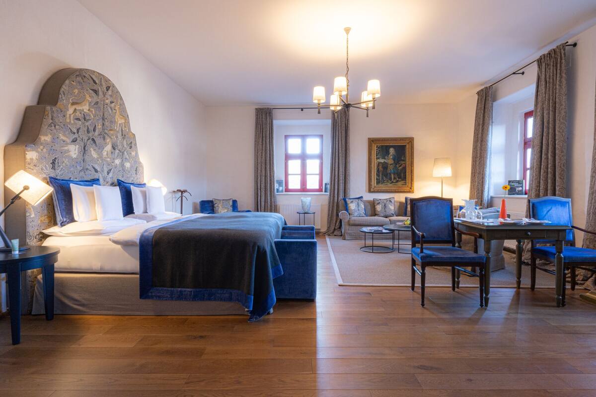 A hotel room with a large bed, a sofa, a dining table with chairs, and a chandelier.