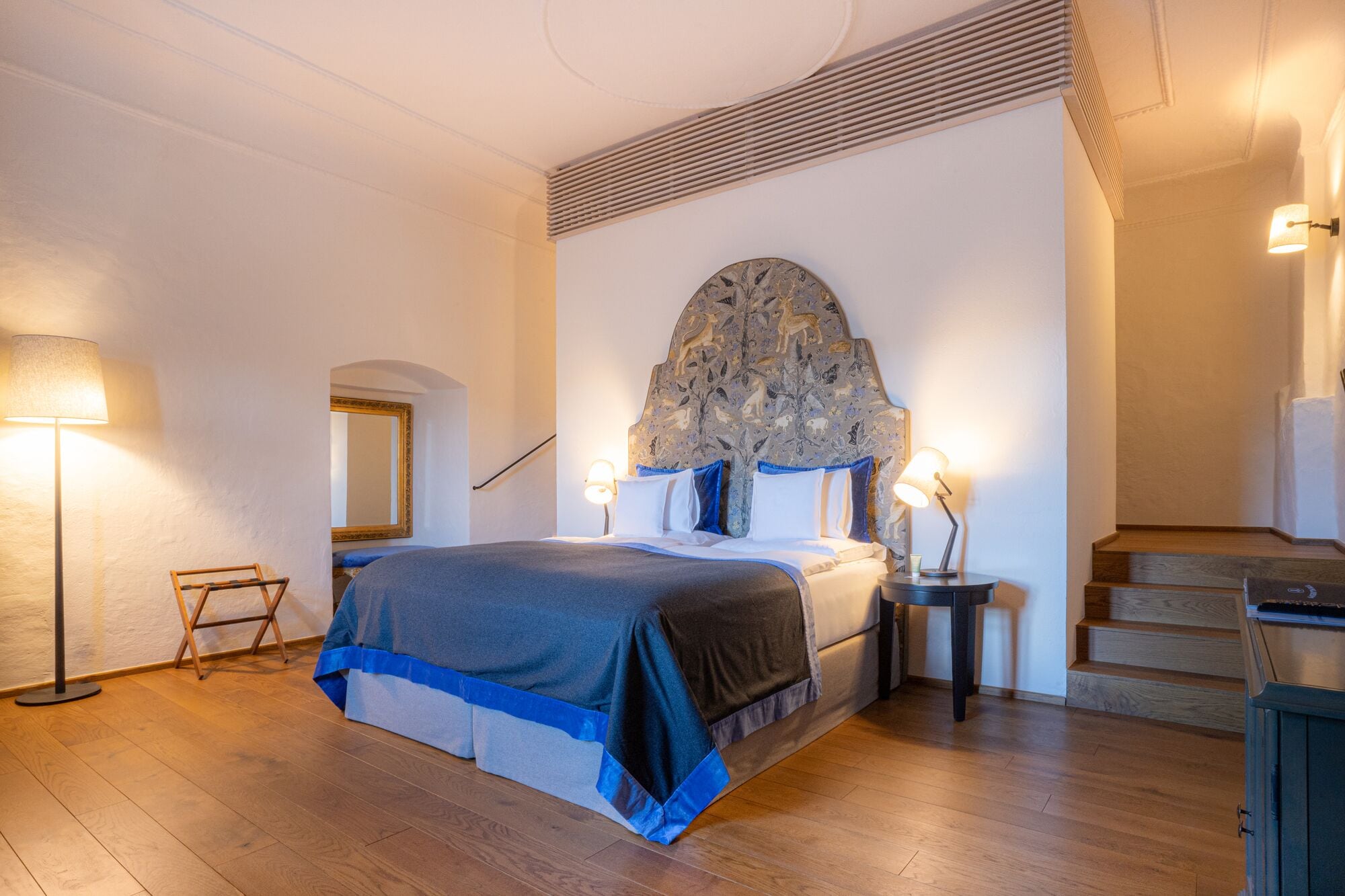 Een slaapkamer met een groot bed, twee bedlampjes, een hoofdeinde met een bloemendecor, een spiegel, en een trap die naar een verdieping leidt.