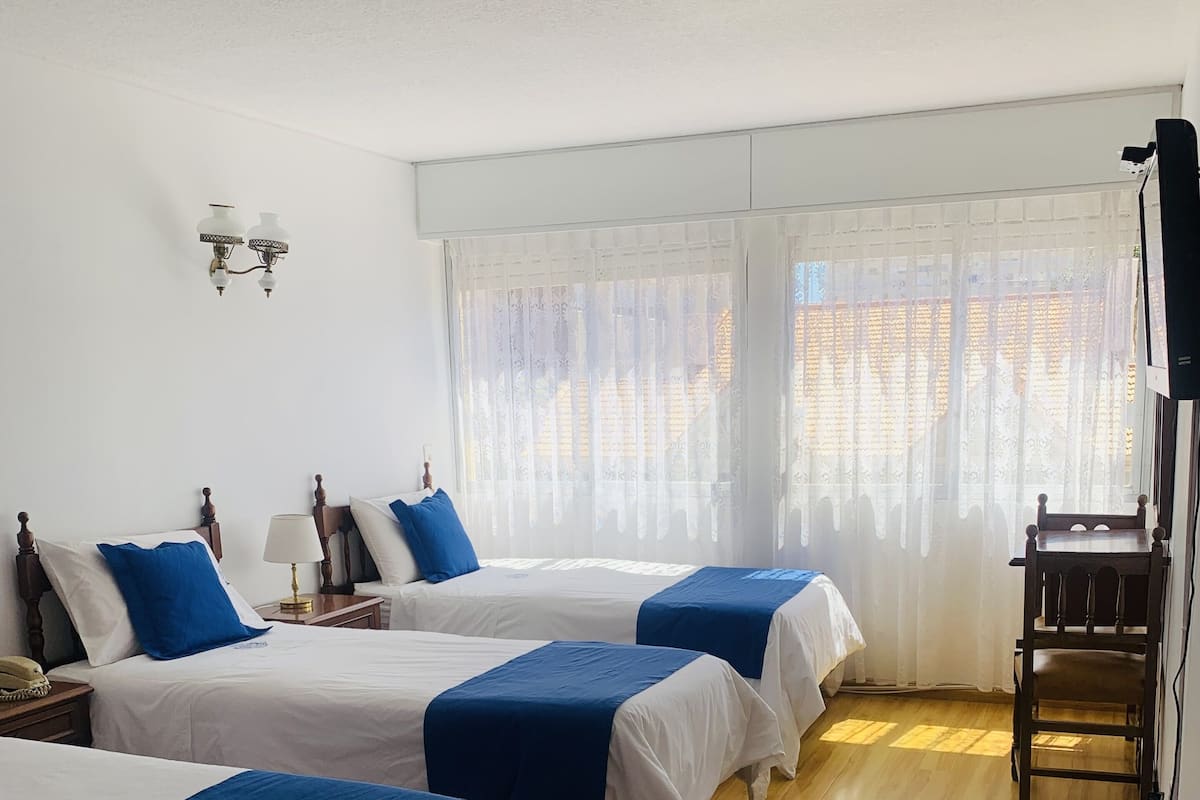 Habitación de hotel con tres camas individuales, suelo de madera, televisor y ventana con cortinas traslúcidas.