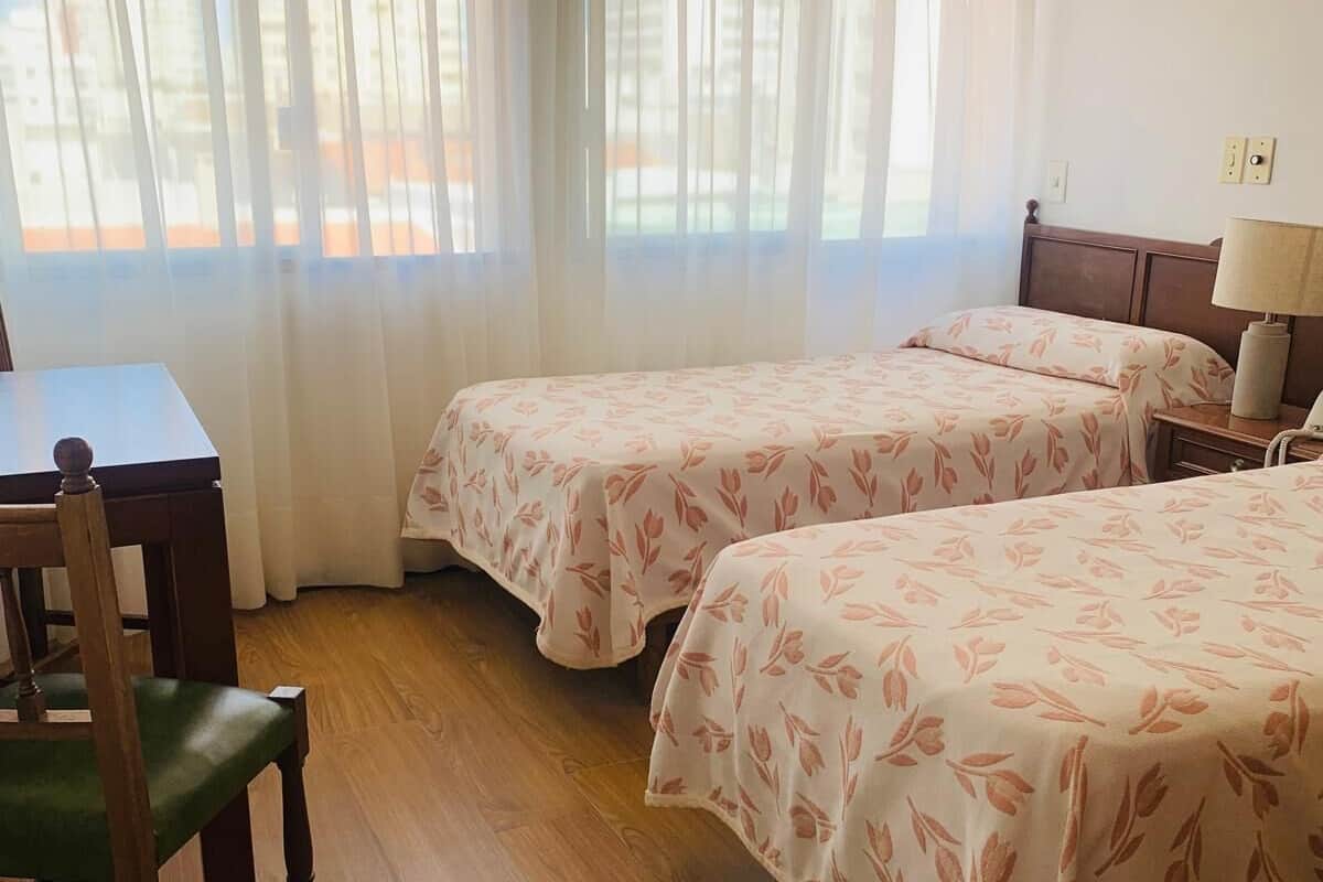 Habitación de hotel con dos camas, una silla de madera, una mesa y una lámpara.