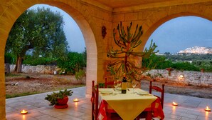 An outdoor dining area with a table set for two, surrounded by stone arches, potted plants, and candles.