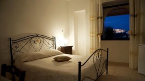 A single bed with a headboard, a bedside table, a window with curtains, and a view of a cityscape at dusk.