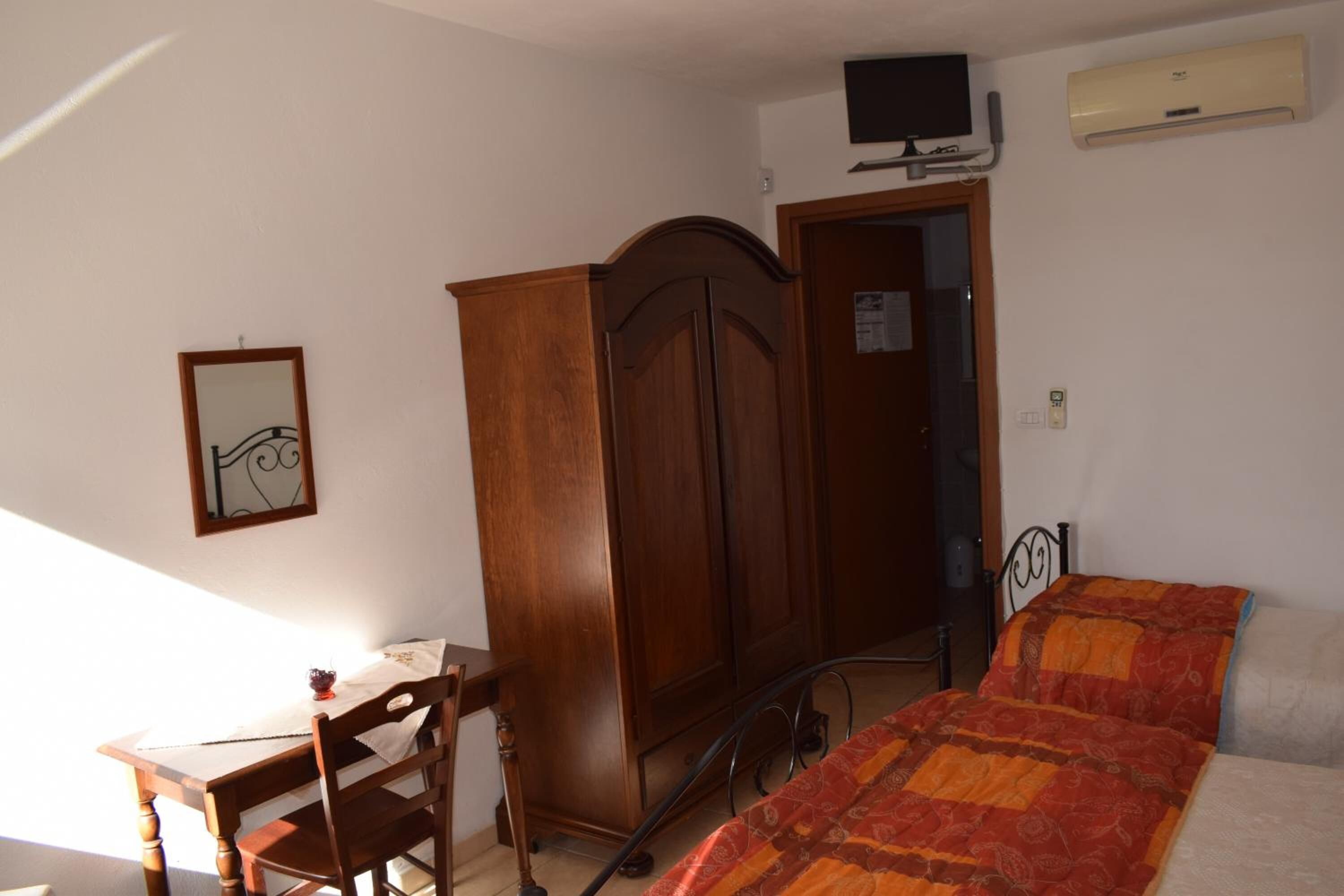 A bedroom with a bed, a wooden wardrobe, a desk with a chair, a television mounted on the wall, and an air conditioning unit.