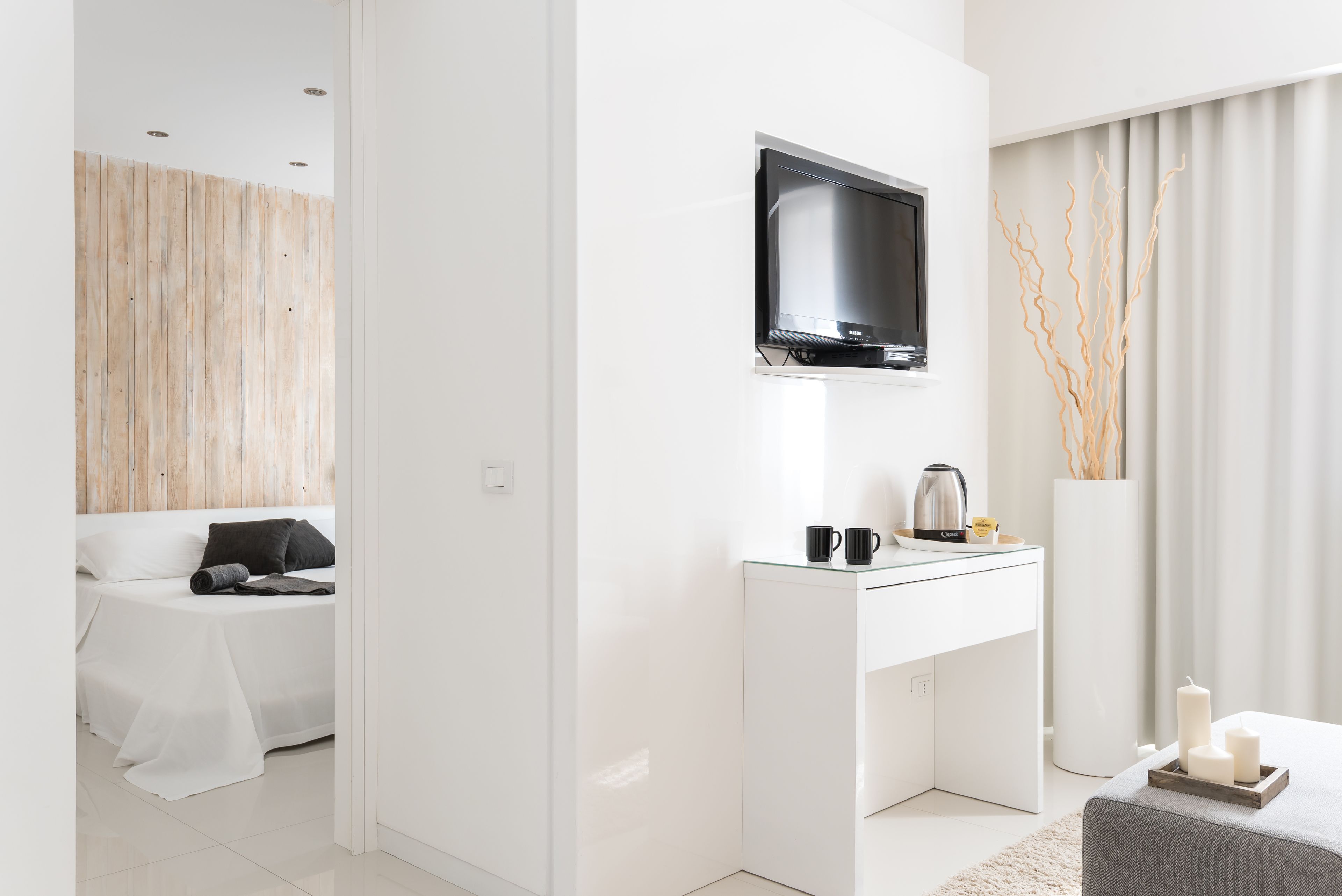 A modern hotel room with a flat-screen TV mounted above a white desk, a bed with white linens, and a large vase with decorative branches.