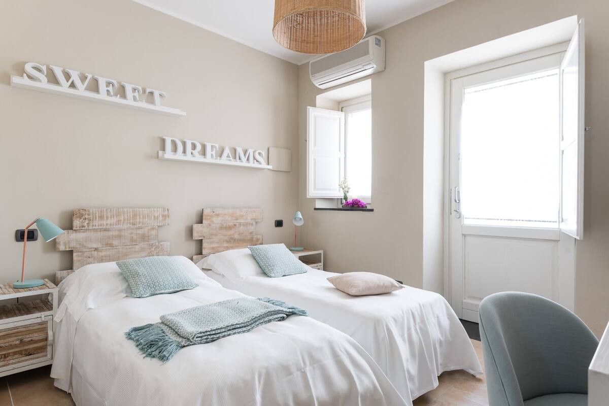 A bedroom with two beds, a wicker chair, a bedside table with a lamp, and a window with a view.