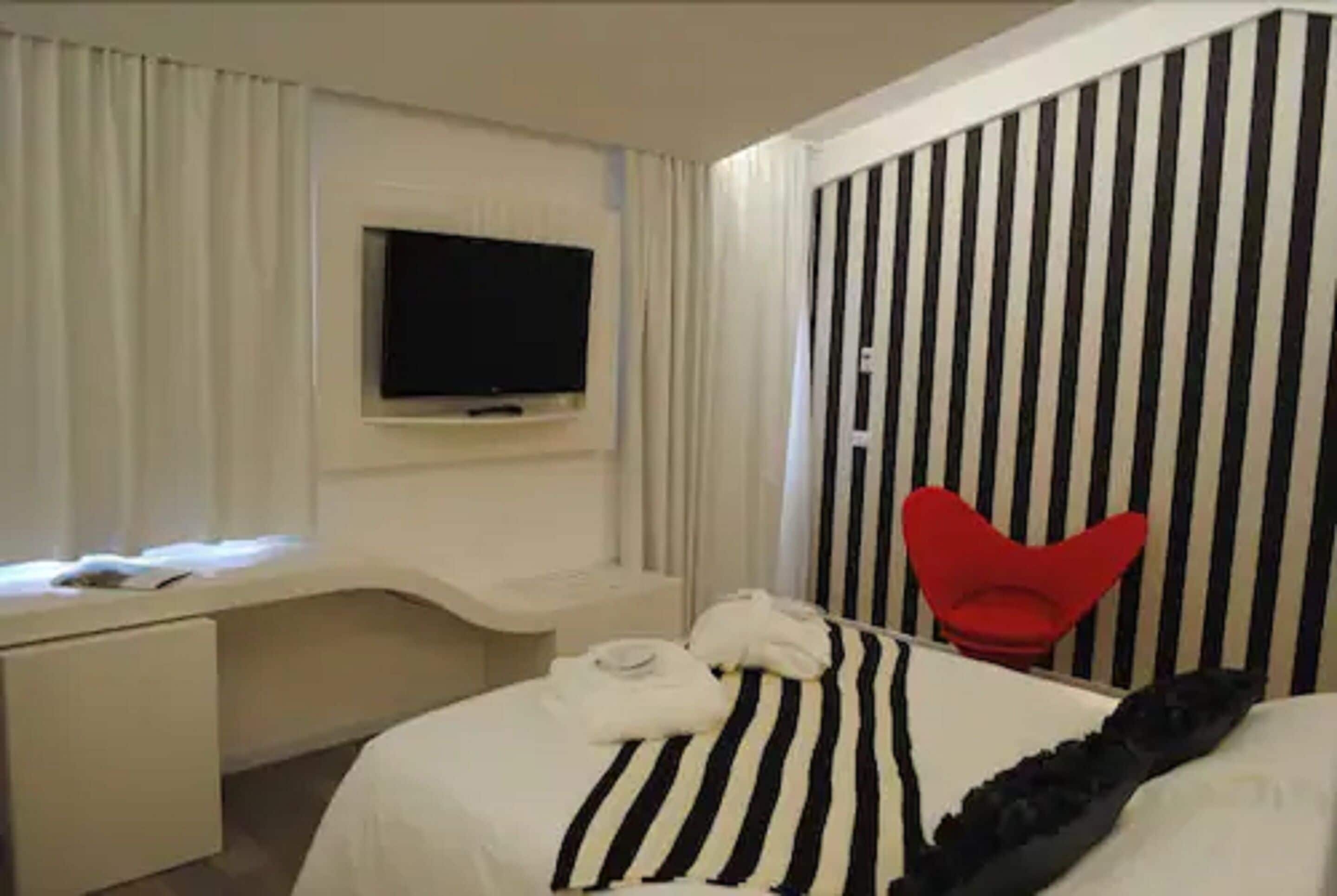 Una habitación de hotel con una cama, un televisor montado en la pared, un escritorio y una silla roja.