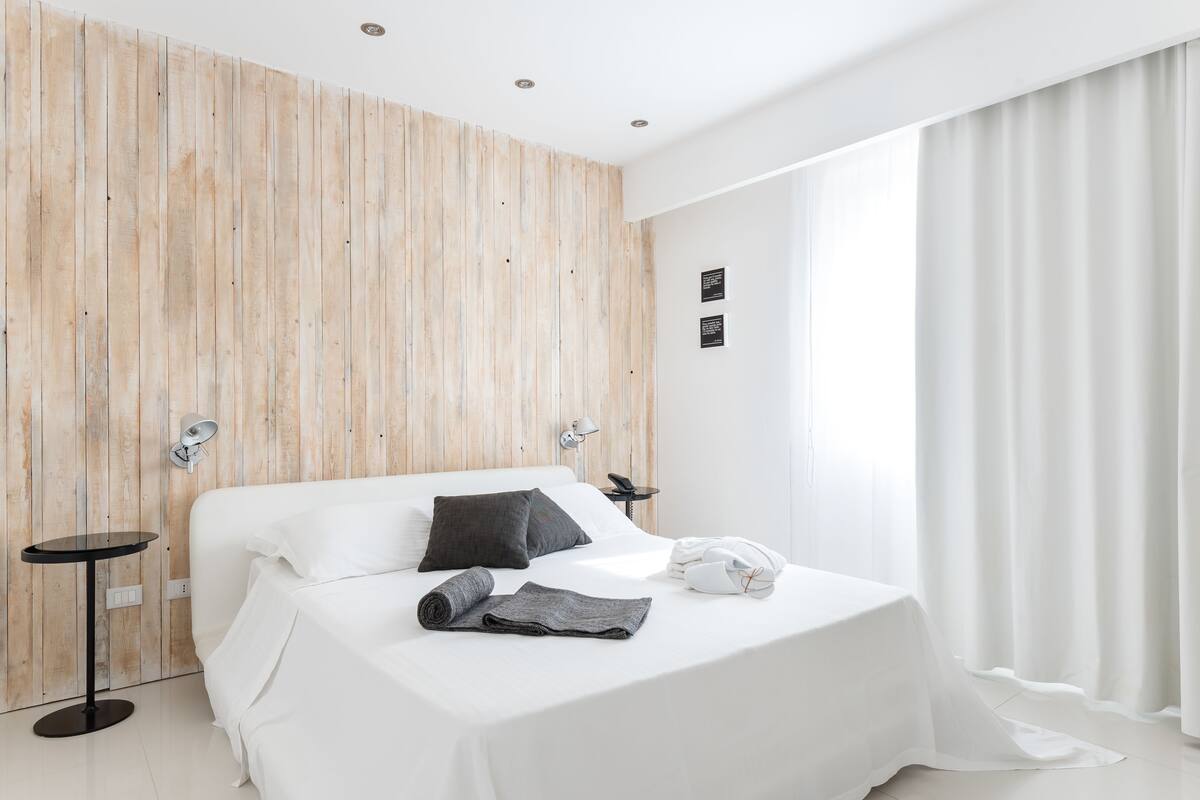 A modern hotel room with a wooden accent wall, a neatly made bed, a small round table, and a bedside lamp.