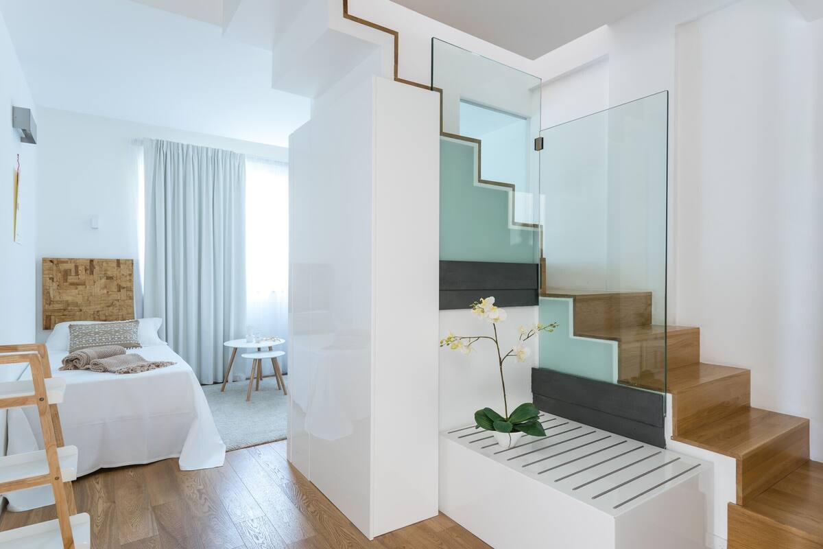 A modern hotel room with a bed, a small table, a chair, a staircase with glass railings, and a potted orchid.