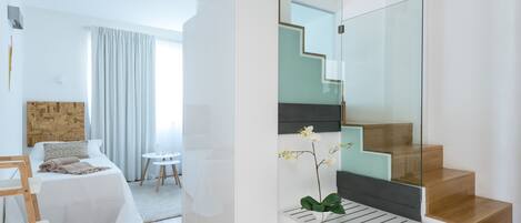 A modern hotel room with a bed, a small table, a chair, a staircase with glass railings, and a potted orchid.