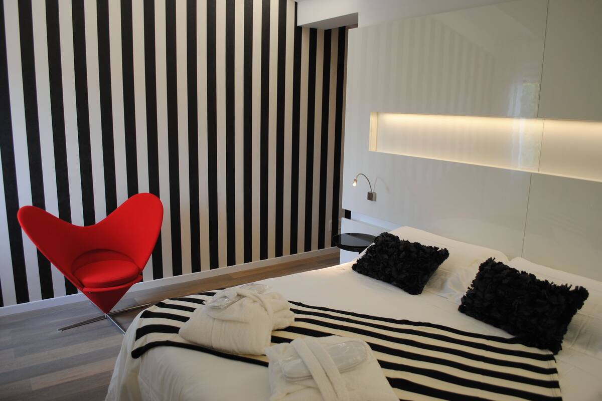 A modern hotel room with a striped wall, a red chair, a bed with black and white bedding, and a bedside table with a lamp.