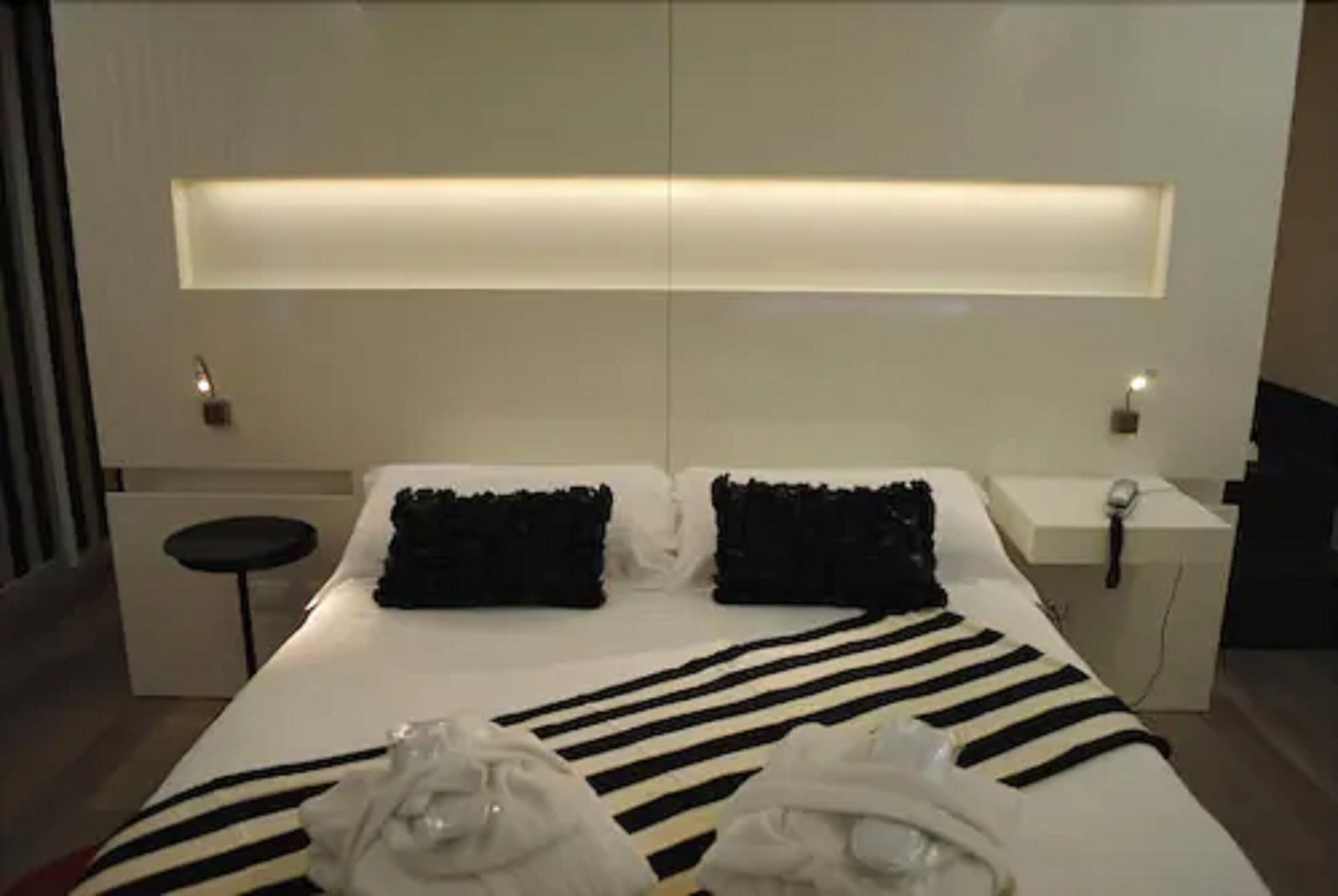 A modern hotel room with a large bed, black and white striped bedding, a bedside table with a lamp, and a small stool.