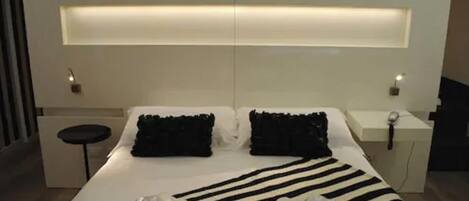 A modern hotel room with a large bed, black and white striped bedding, a bedside table with a lamp, and a small stool.