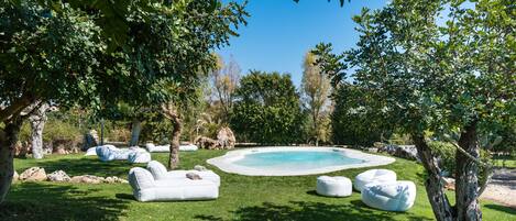 Zona de piscina con sillones blancos y pufs, rodeada de árboles y rocas.