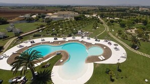 Vista aérea de un complejo turístico con una piscina grande de forma singular, rodeada de áreas de descanso y palmeras.