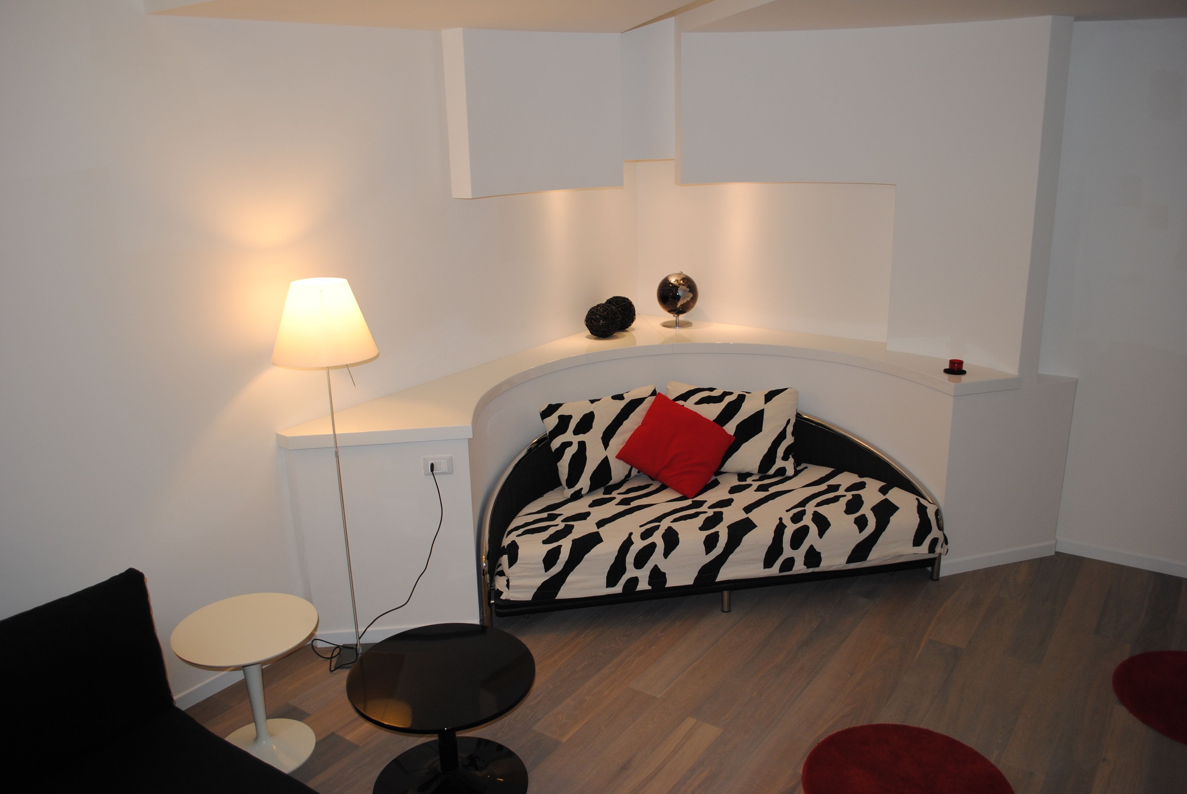 Ein modernes Schlafzimmer mit Schlafsofa, einem kleinen runden Tisch, einer Lampe und einem roten Zierkissen.