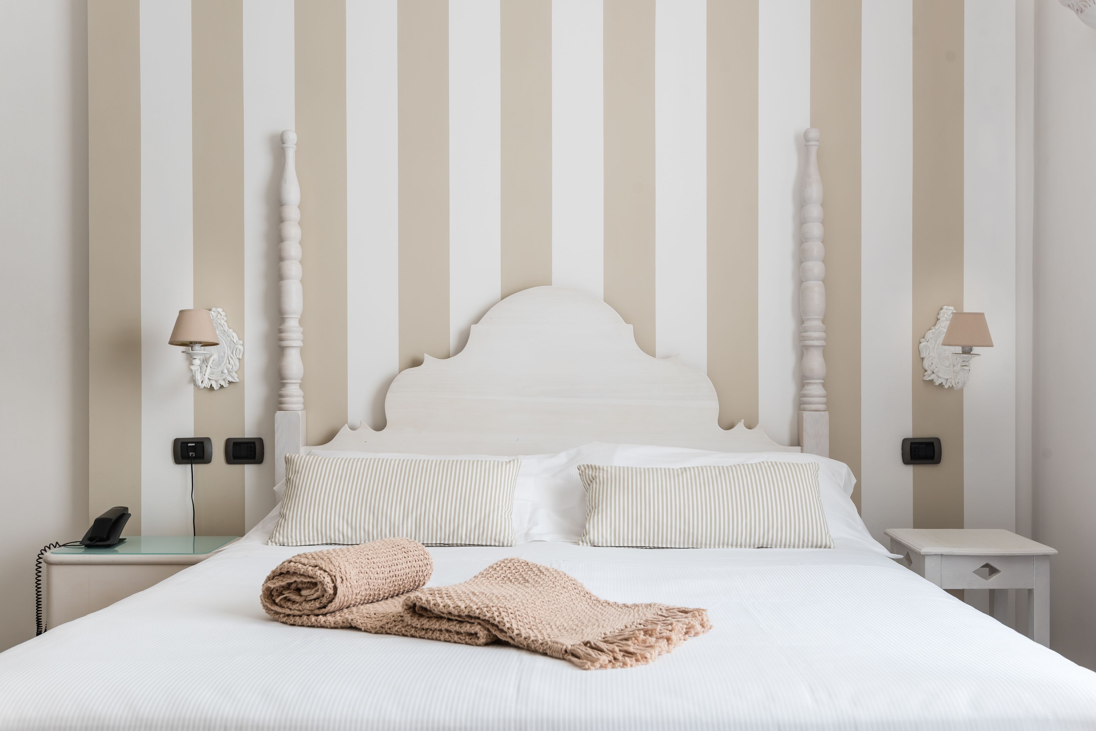 A neatly made bed with a striped pillow, a rolled towel, and a white headboard against a striped wall.