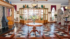 Una stanza con pavimento a scacchi, un tavolo centrale con un vaso di fiori, un camino in pietra e un mobile in legno con oggetti decorativi.