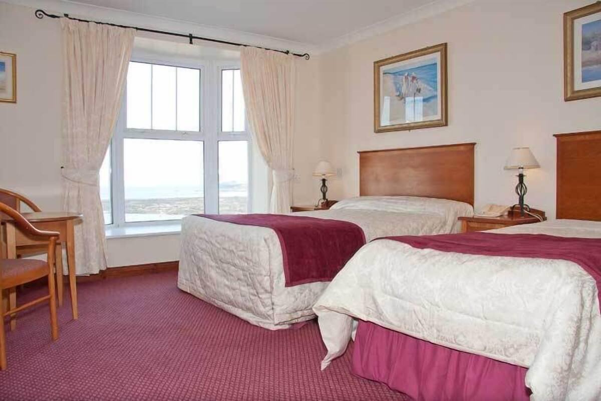 Una habitación de hotel con dos camas, una silla de madera, una ventana con cortinas y un cuadro en la pared.